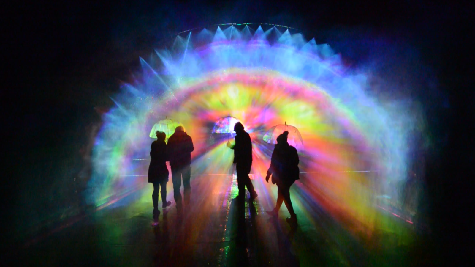 A trio of figures silhoutted by rainbow lights at Light Up Poole 